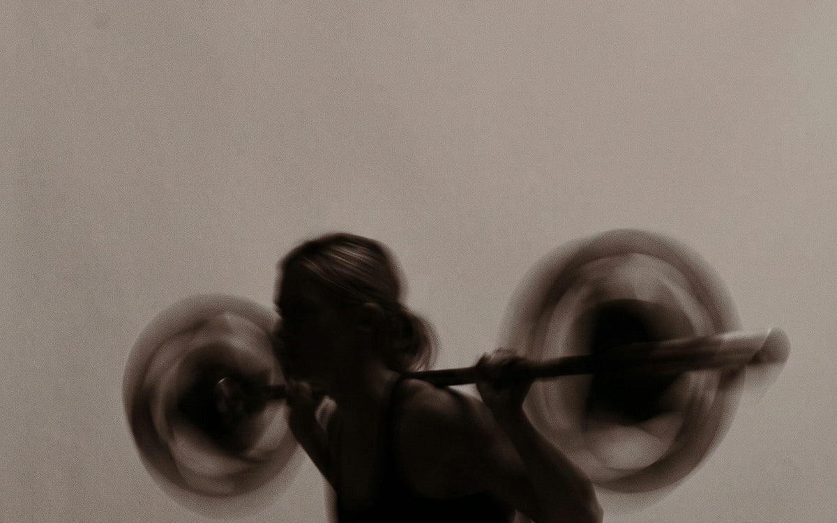A lifter squatting under a barbell, motion-blurred against a cream wall