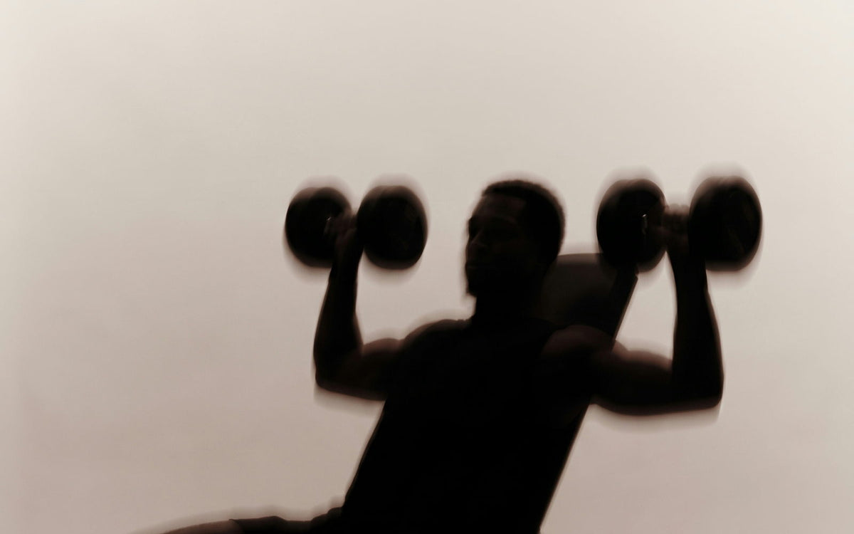 A seated figure pressing dumbbells overhead, motion-blurred against a cream wall