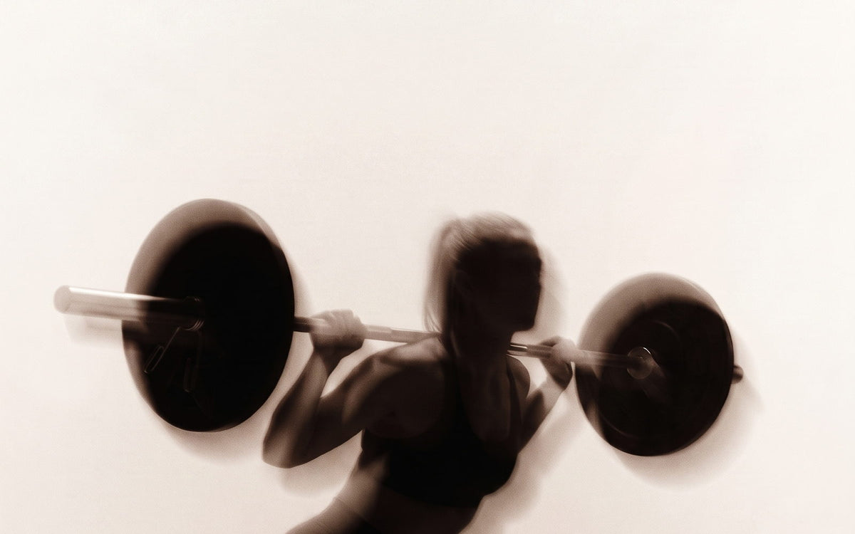 A lifter squatting under a loaded barbell, motion-blurred against a cream wall